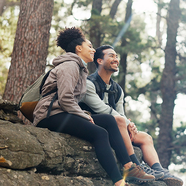 Nature, hiking and a couple relax on adventure trail in forest and sitting on a rock. Health, happy man and woman pointing at natural landscape while relaxing in woods with trees, fitness and freedom